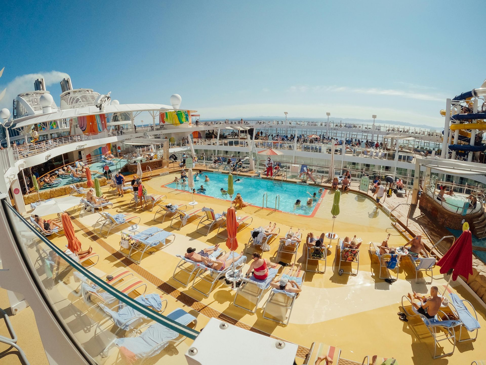High-angle view of a cruise ship pool deck with sun loungers, a swimming pool, and water slides under a bright blue sky.