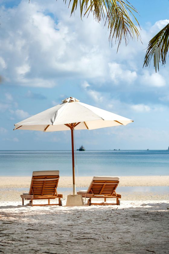 Two empty wooden lounge chairs sit under a white umbrella on a sunny, sandy beach by the ocean.