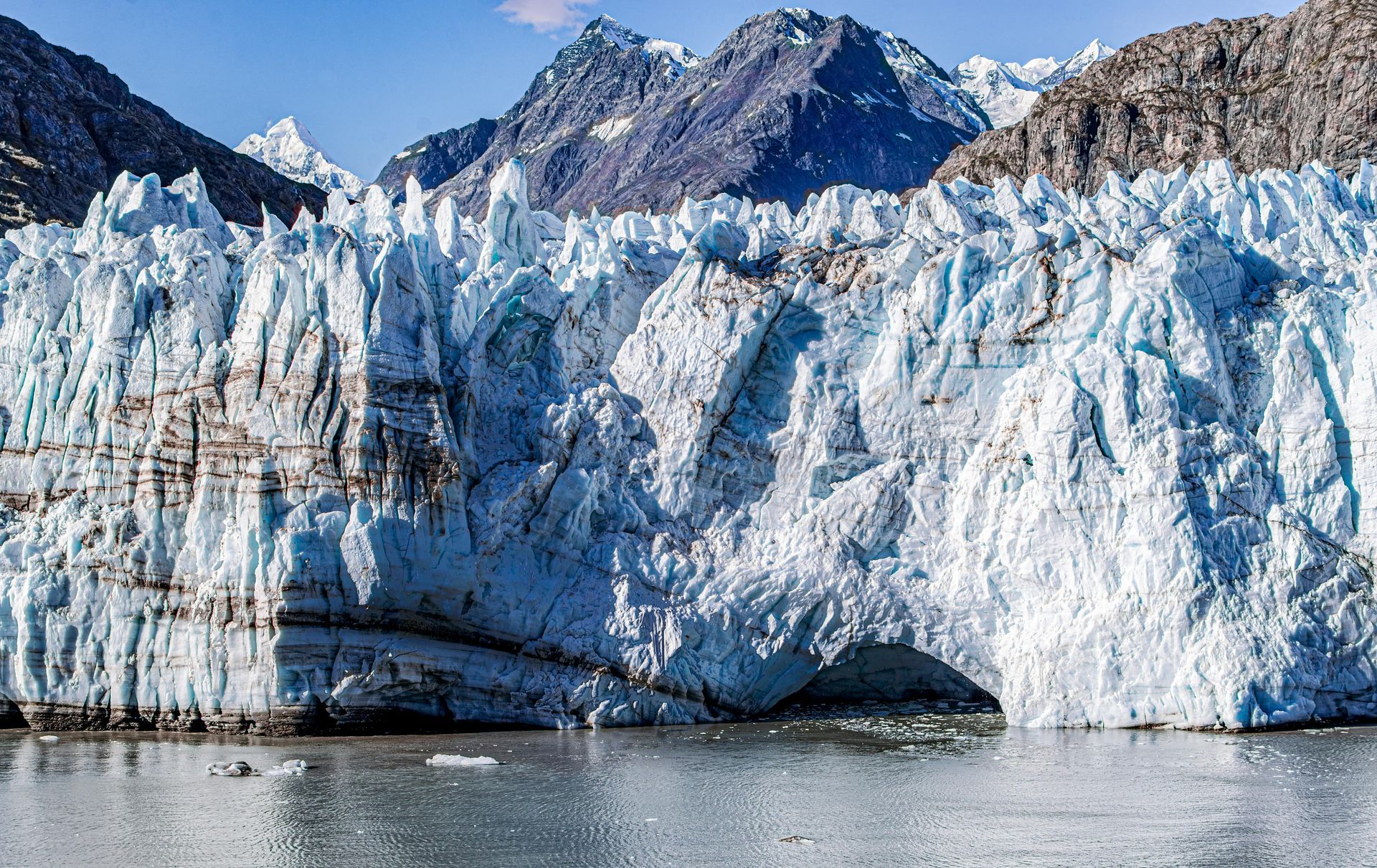 Jagged blue and white glacier ice rises above calm, dark water, set against a backdrop of rugged mountain peaks.