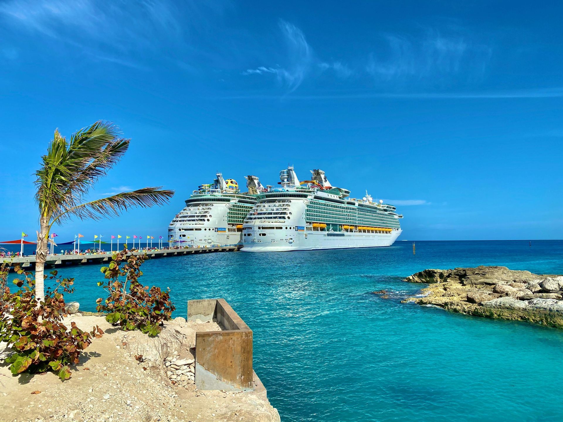 Two large cruise ships are docked at a pier in crystal-clear turquoise waters under a bright blue sky.