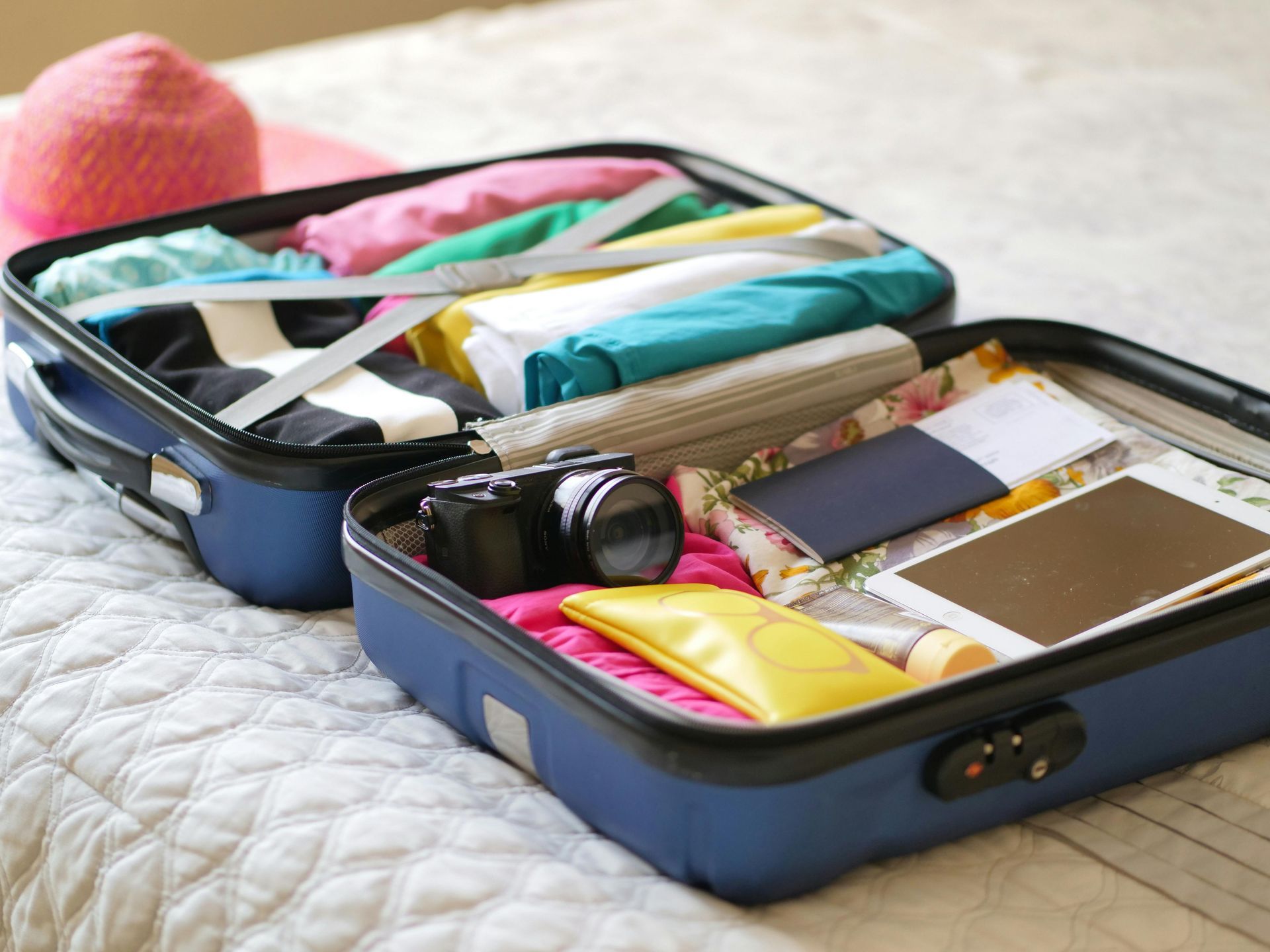 An open blue suitcase on a bed, neatly packed with folded colorful clothes, a camera, a tablet, and travel documents.