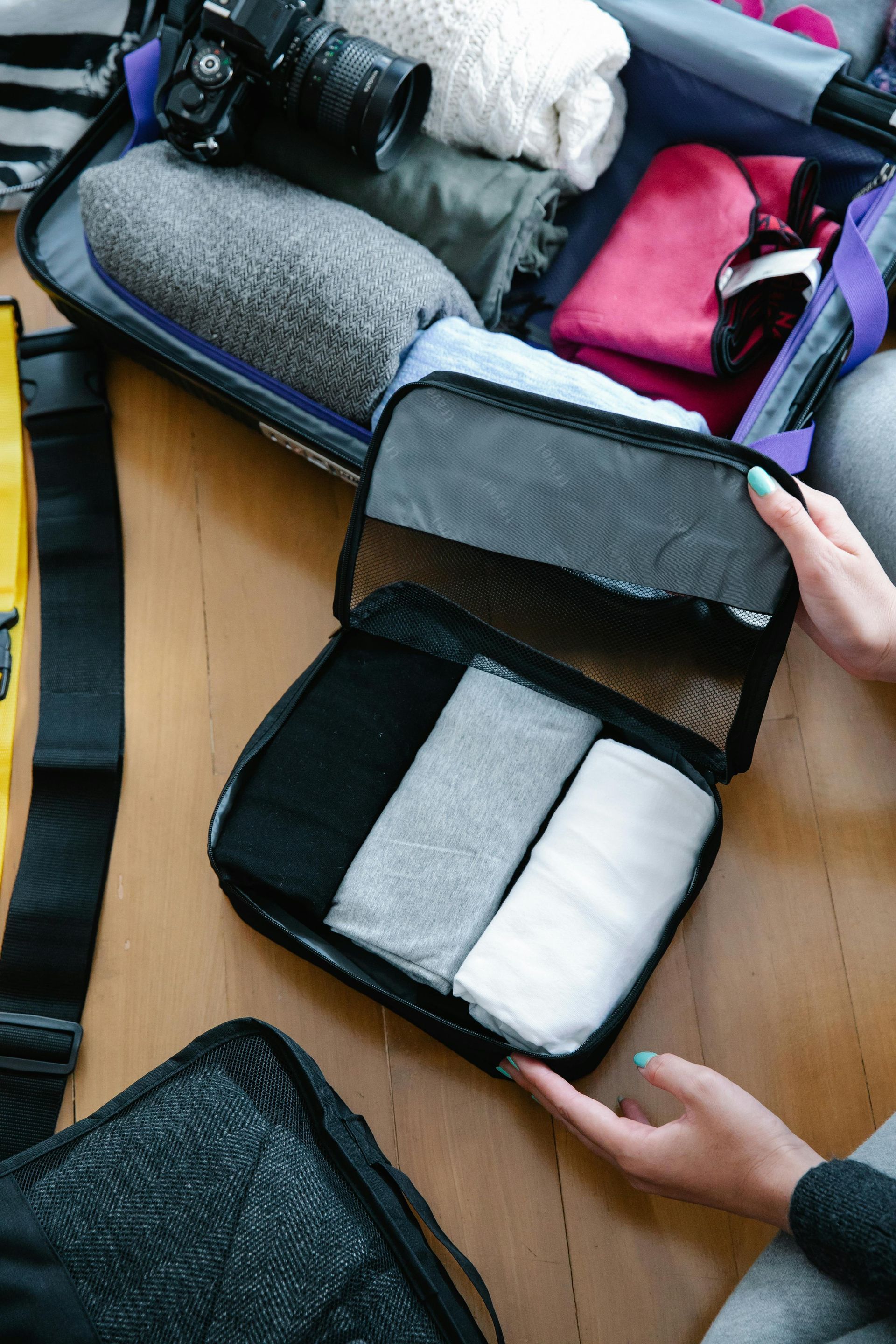 Hands packing clothes into a zippered mesh travel organizer cube next to an open suitcase on a wooden floor.