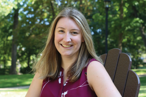 A smiling person with long blonde hair, wearing a maroon patterned sleeveless top, sits in a park chair.