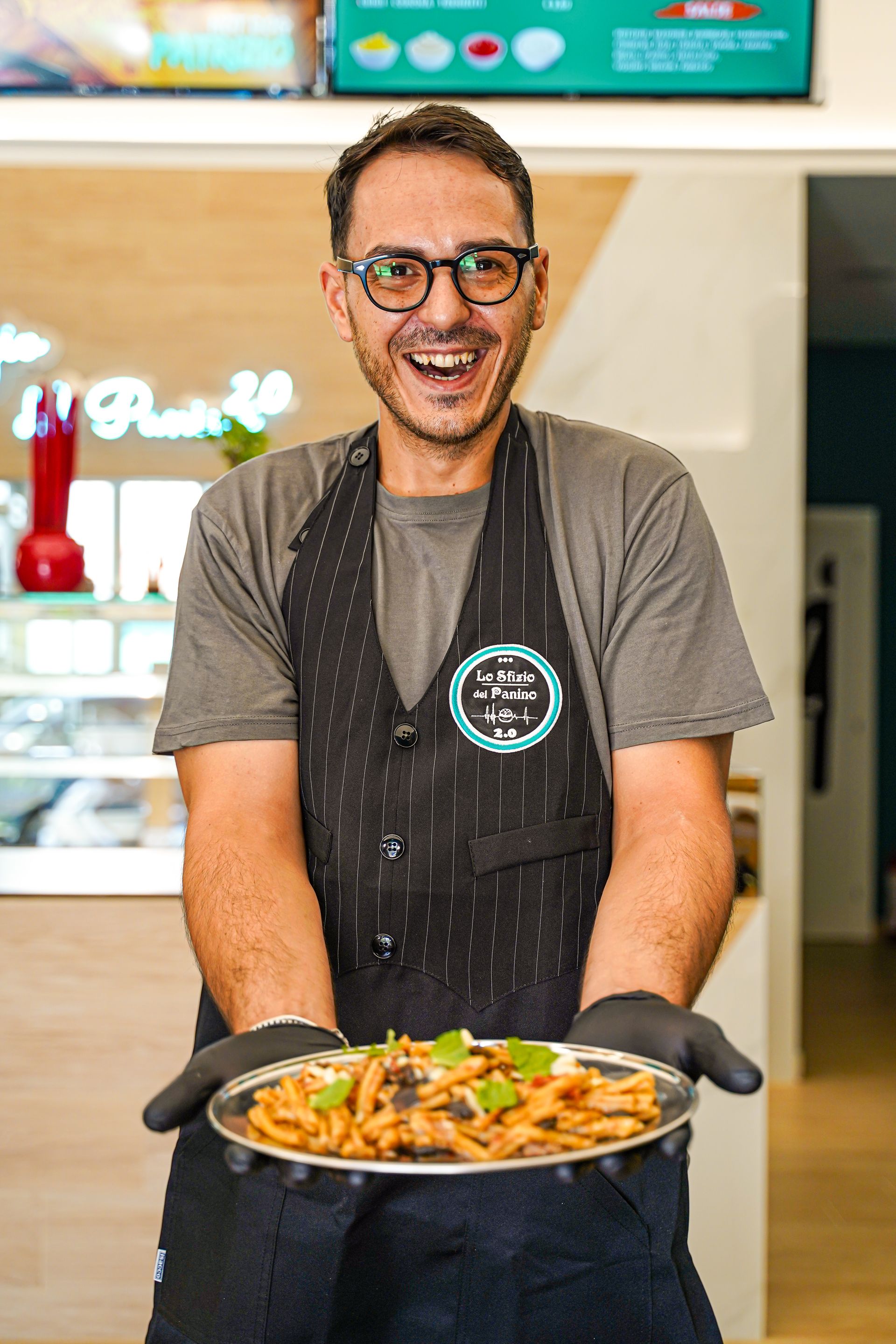 Uomo sorridente con in mano un piatto di pasta. Indossa occhiali, grembiule e guanti neri in un ristorante.