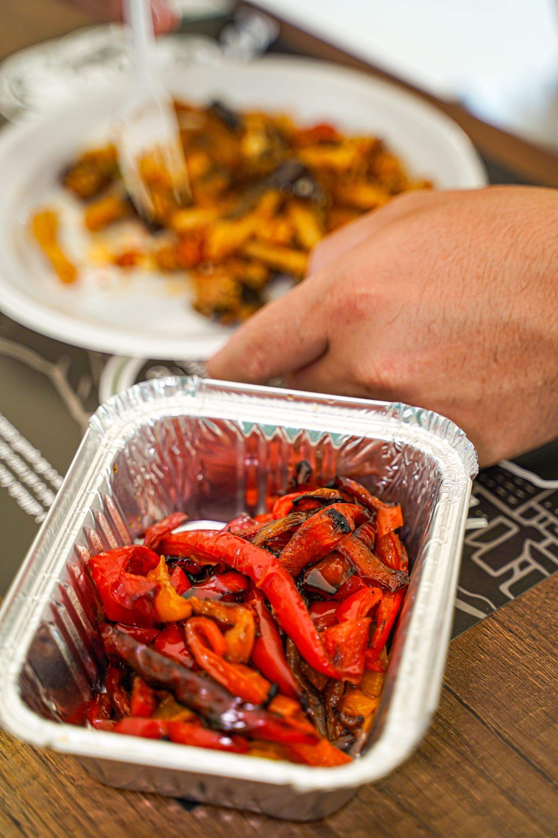 Peperoni rossi arrostiti in un contenitore di alluminio, sullo sfondo una persona che prende un piatto di pasta.