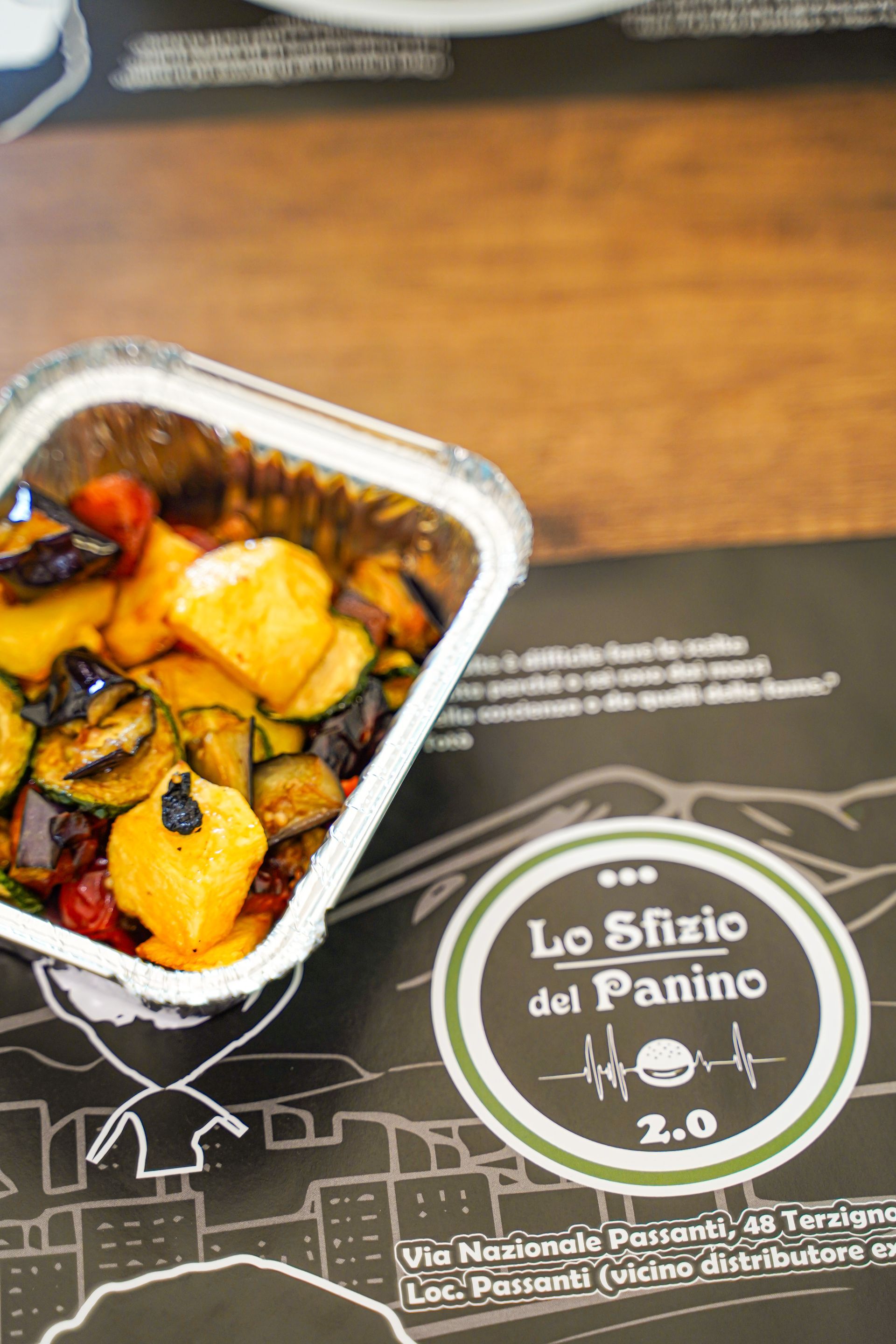 Verdure arrostite in un contenitore di alluminio su una tovaglietta da ristorante con il logo.