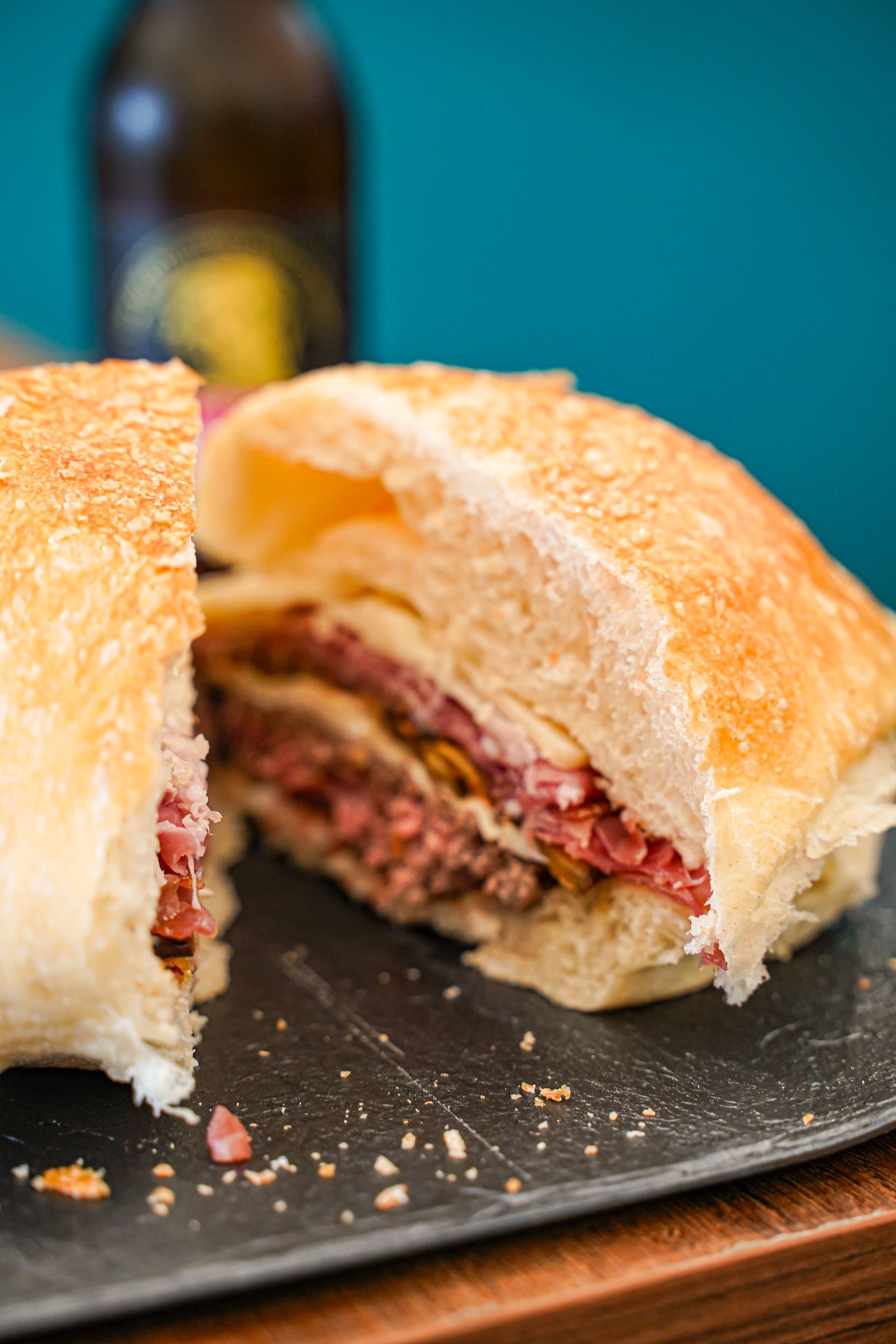 Un panino affettato ripieno di carne, formaggio e condimenti, su un tagliere nero, con una bottiglia di birra sullo sfondo.