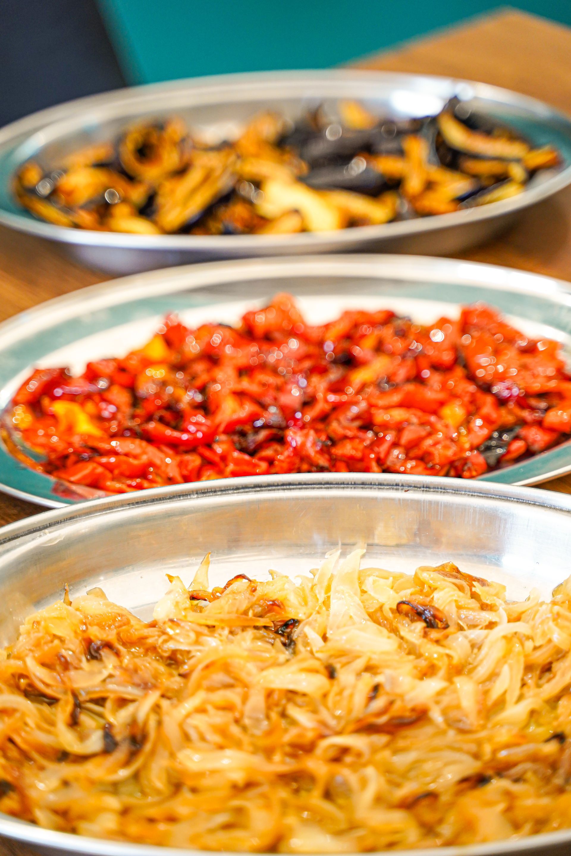 Tre vassoi d'argento con verdure cotte: melanzane, peperoni rossi e cipolle.