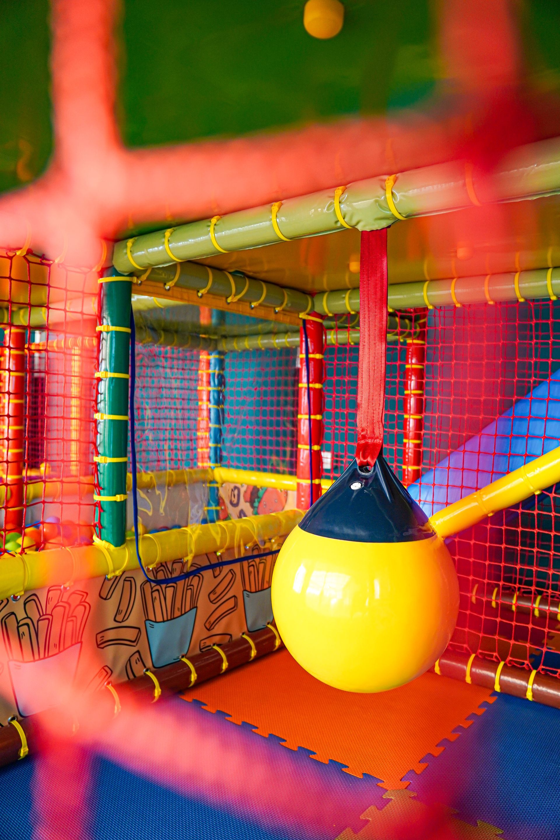 Parco giochi al coperto con palla gialla sospesa, tappetini colorati e rete.