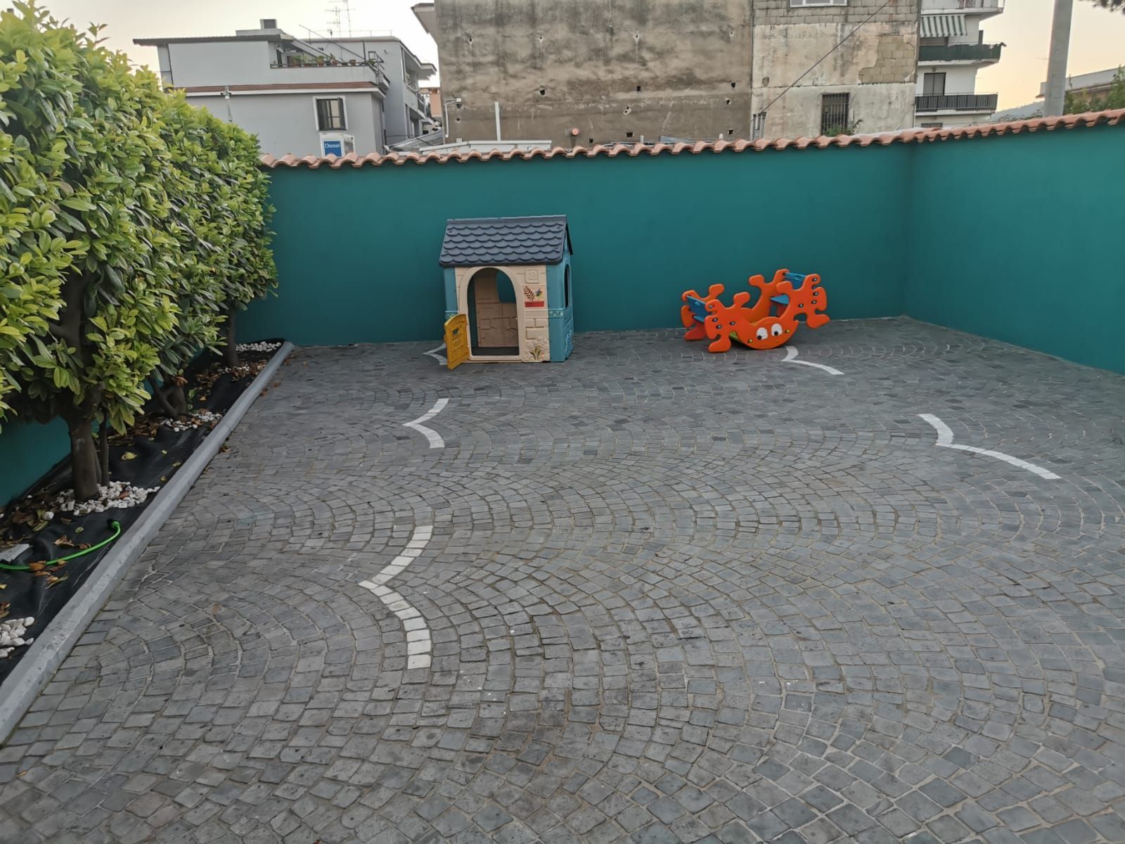 Parco giochi con un muro color verde acqua, ciottoli grigi, una piccola casetta e un giocattolo a dondolo.