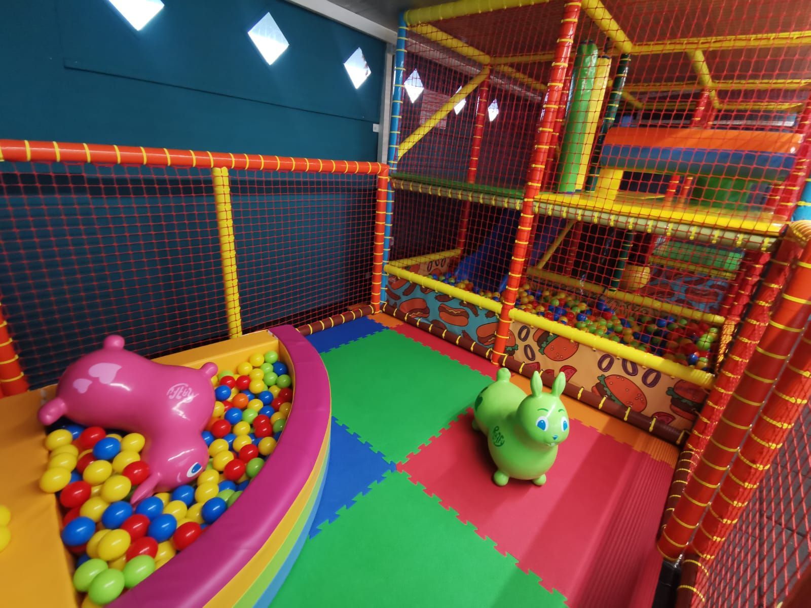 Area giochi interna: piscina di palline, strutture per arrampicarsi, tappeti colorati e giocattoli gonfiabili per bambini.