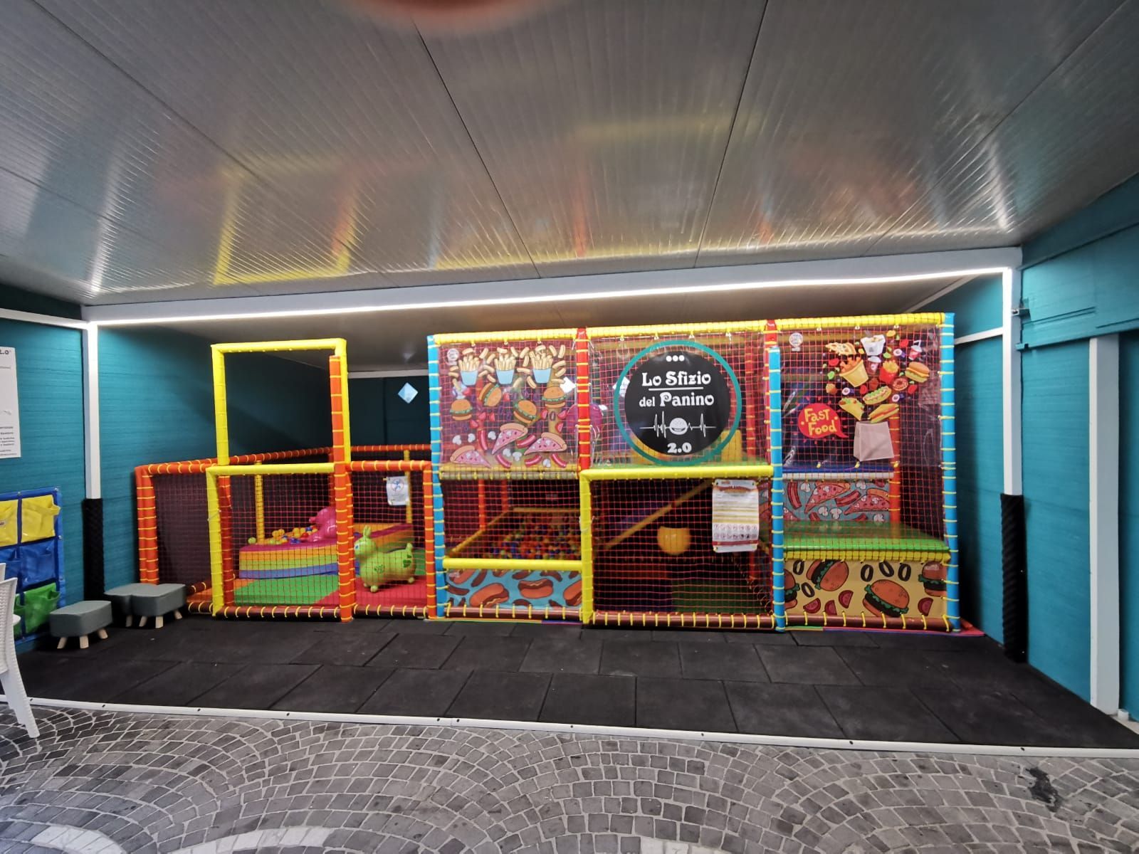 Area giochi interna per bambini con strutture morbide e colorate e piscina di palline all'interno di una struttura con pareti blu.