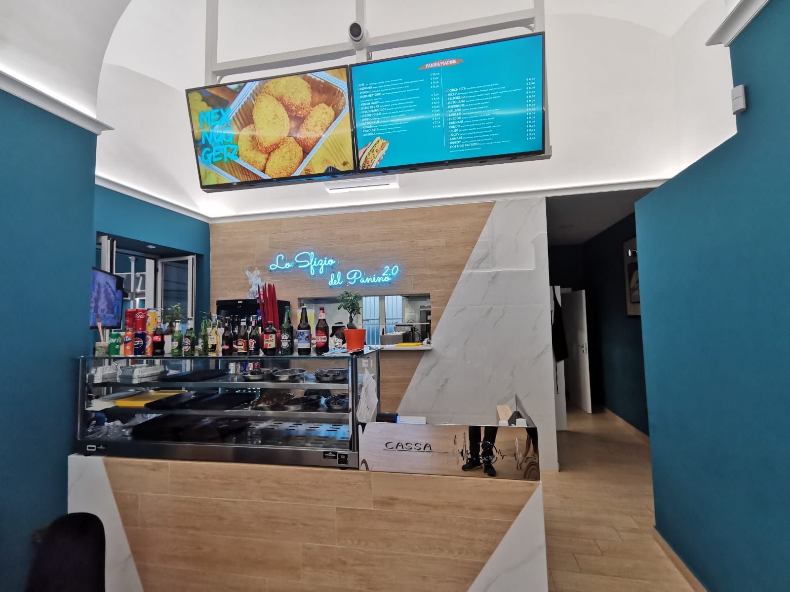 Interno di un negozio di alimentari con bancone, espositore per il menù e area salotto, caratterizzato da decorazioni in turchese e bianco.