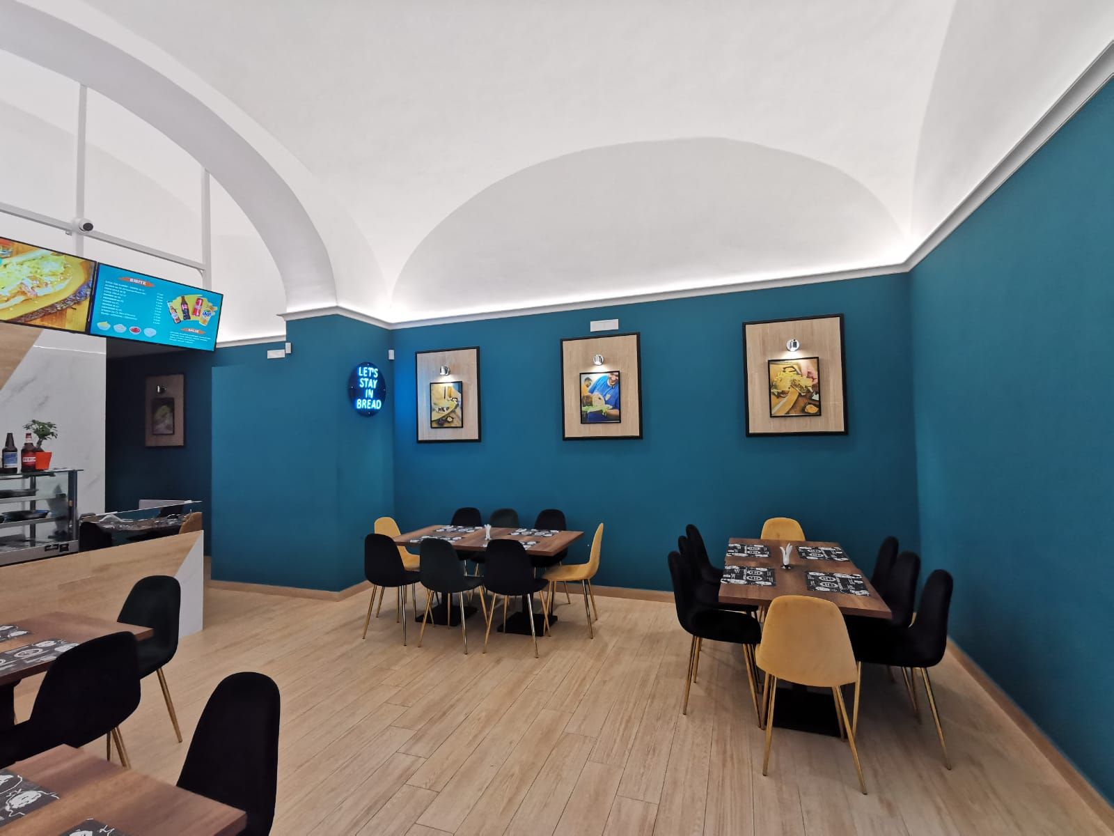 Interno del ristorante con pareti color verde acqua, soffitto bianco ad arco, tavoli in legno, sedie nere e gialle e opere d'arte incorniciate.