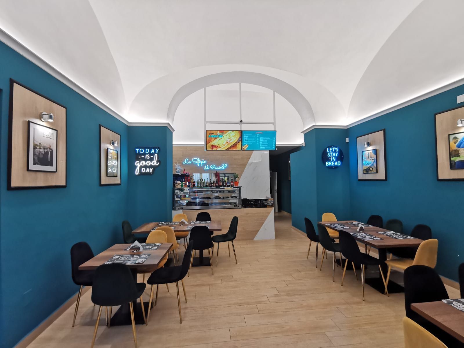 Interno di un ristorante con pareti color verde acqua, sedie gialle e tavoli in legno. Sono visibili un'insegna al neon e un bancone.