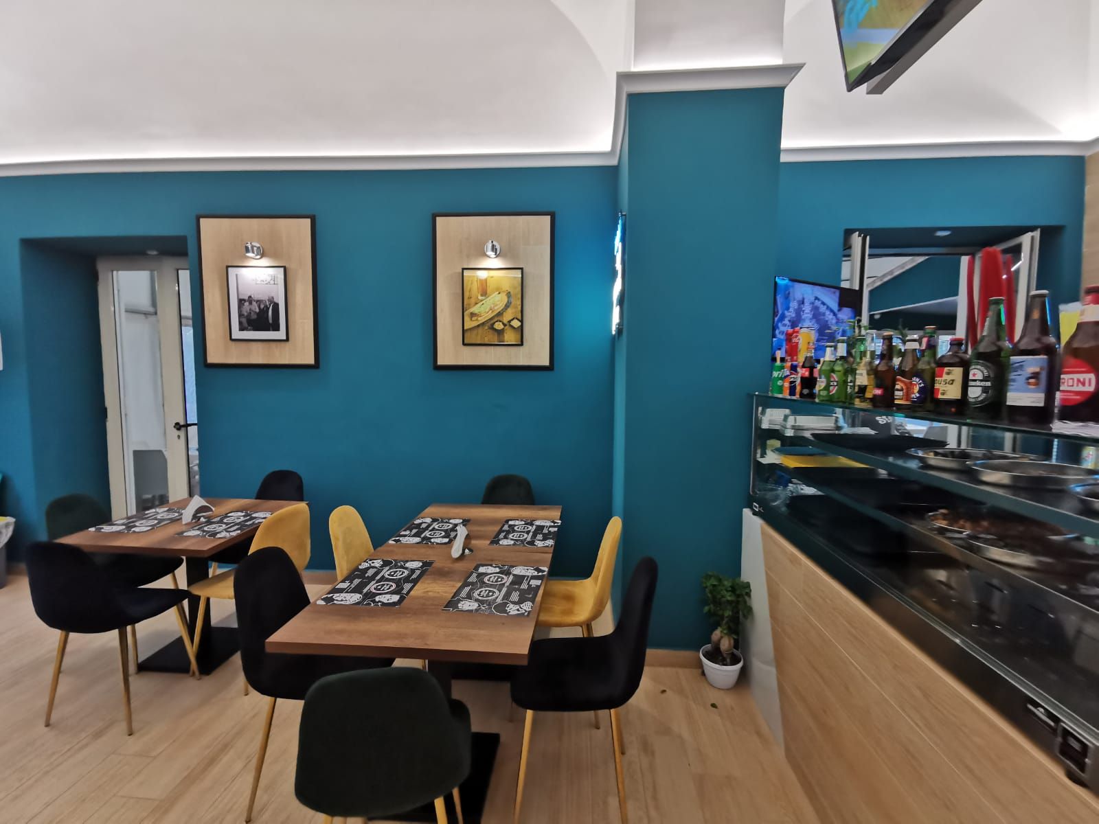 Interno del ristorante con pareti color verde acqua, tavoli con sedie, opere d'arte e un'area bar.