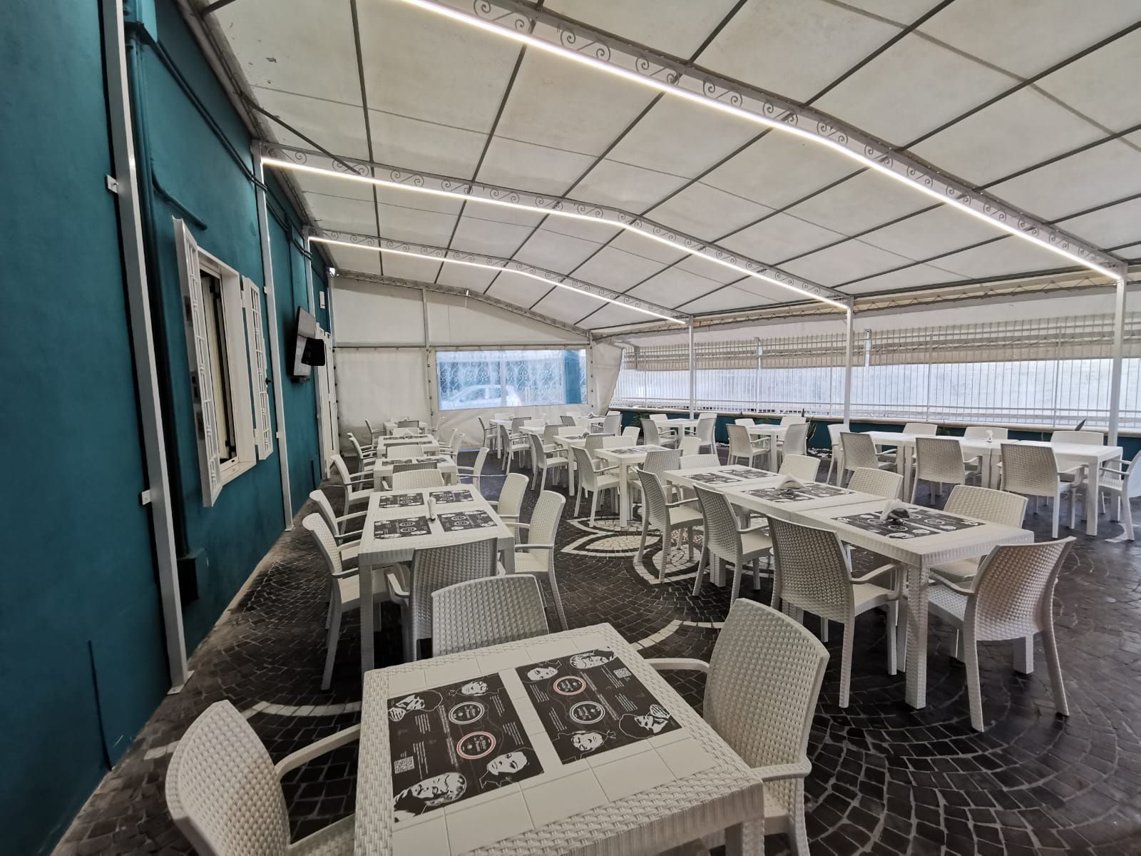 Patio del ristorante con tavoli e sedie bianchi sotto una tettoia bianca; parete dell'edificio color verde acqua.