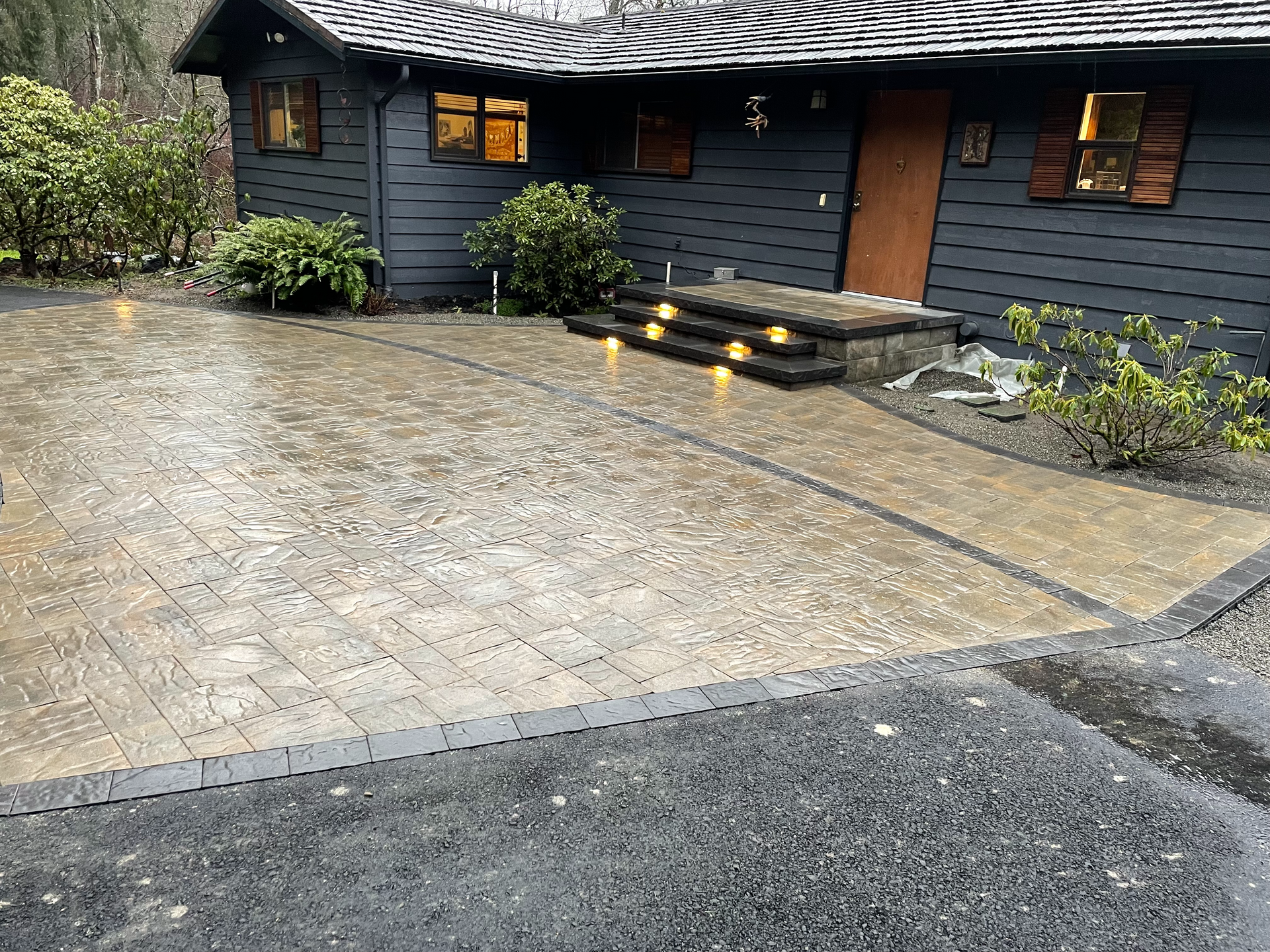 Dark gray house with brick-like driveway and walkway. Rain is falling. Lit steps lead to the front door.
