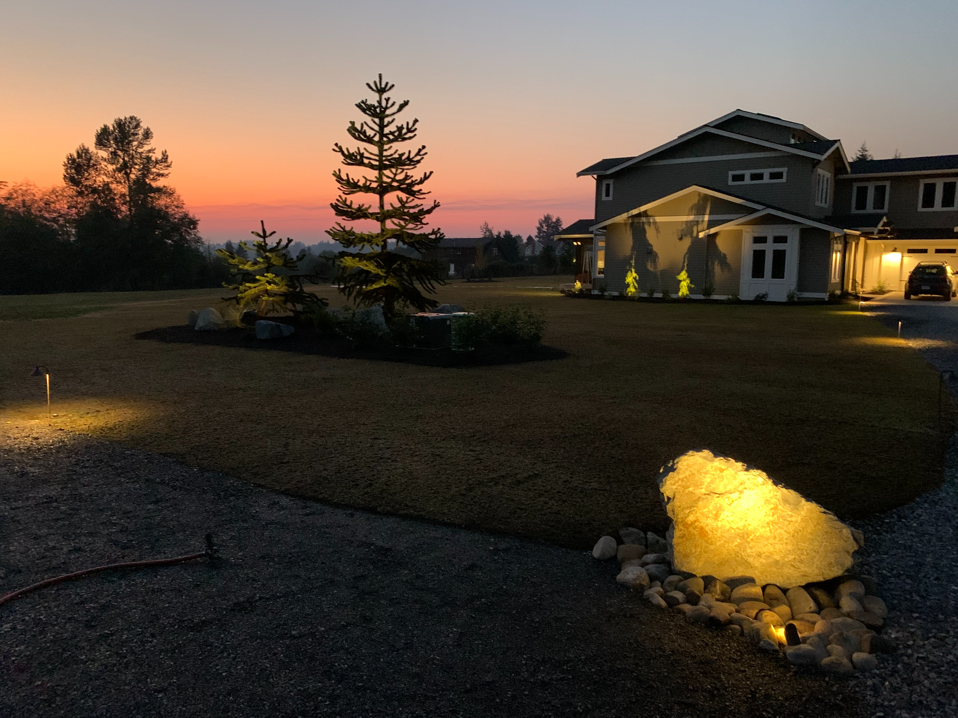 Sunset over a large house with a well-lit yard, gravel driveway, and a decorative illuminated rock.