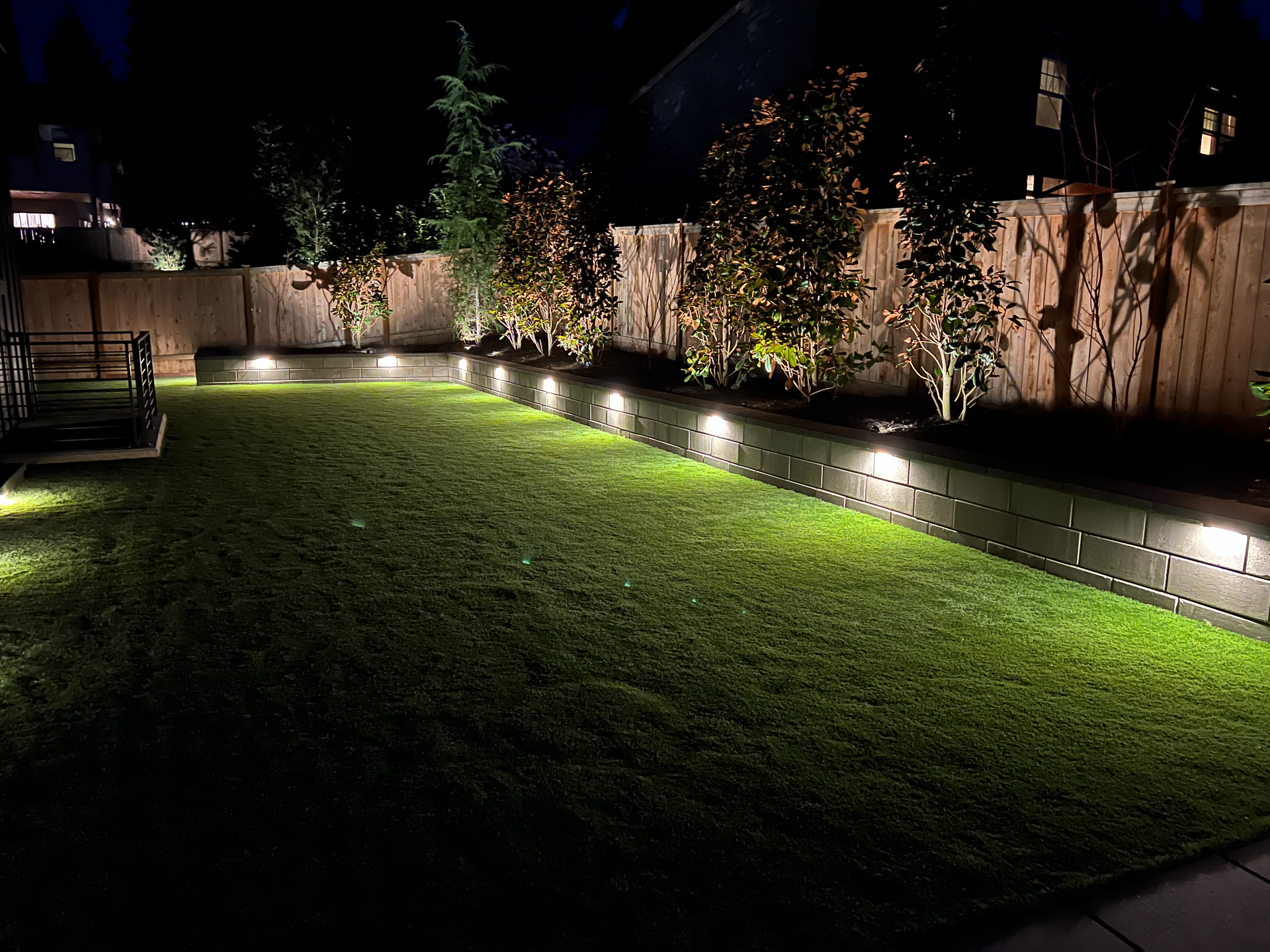 Nighttime view of a backyard with illuminated retaining wall, grass, trees, and fence.