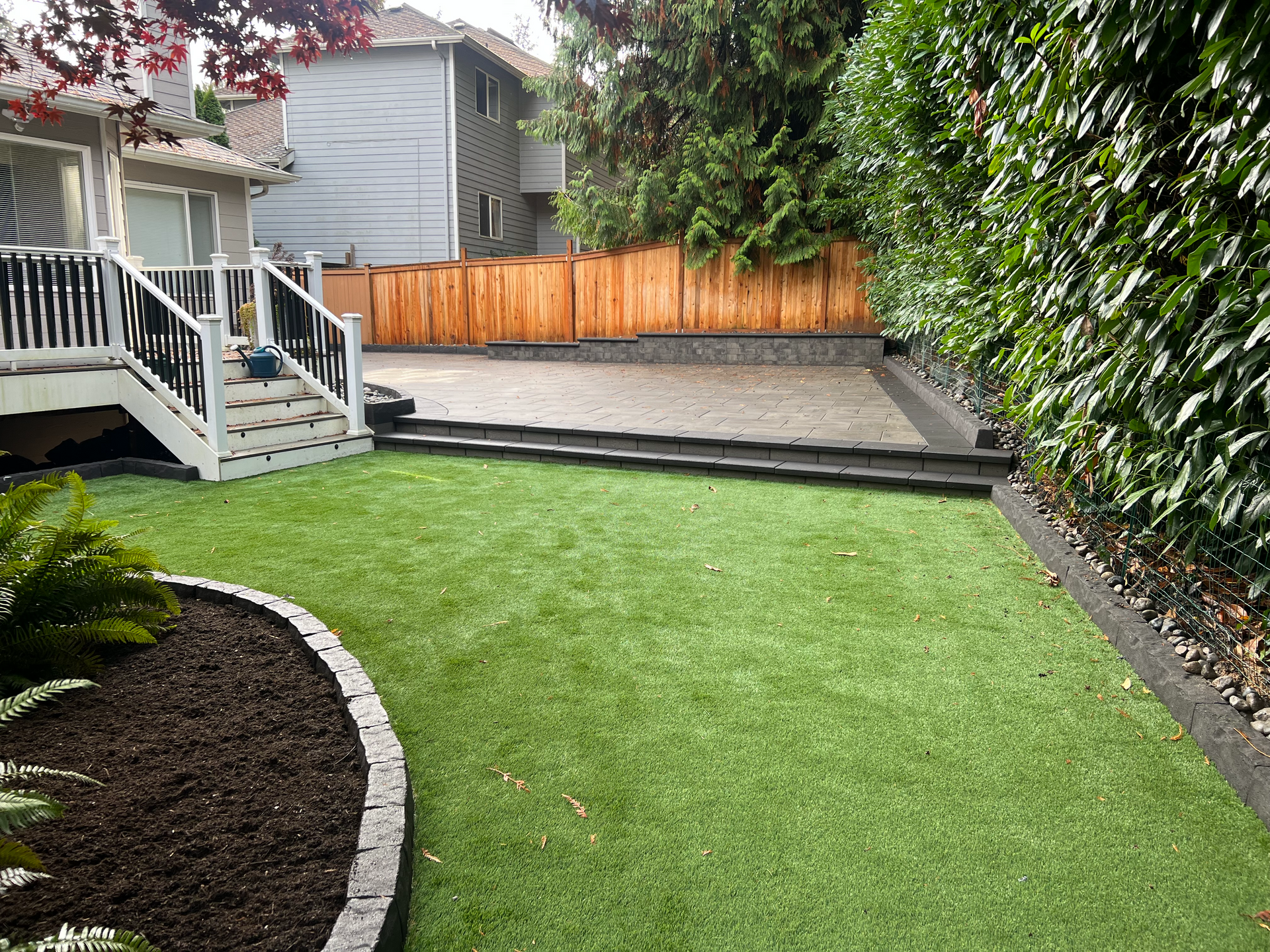 Backyard with green turf, stone patio, wooden fence, and a two-story home.