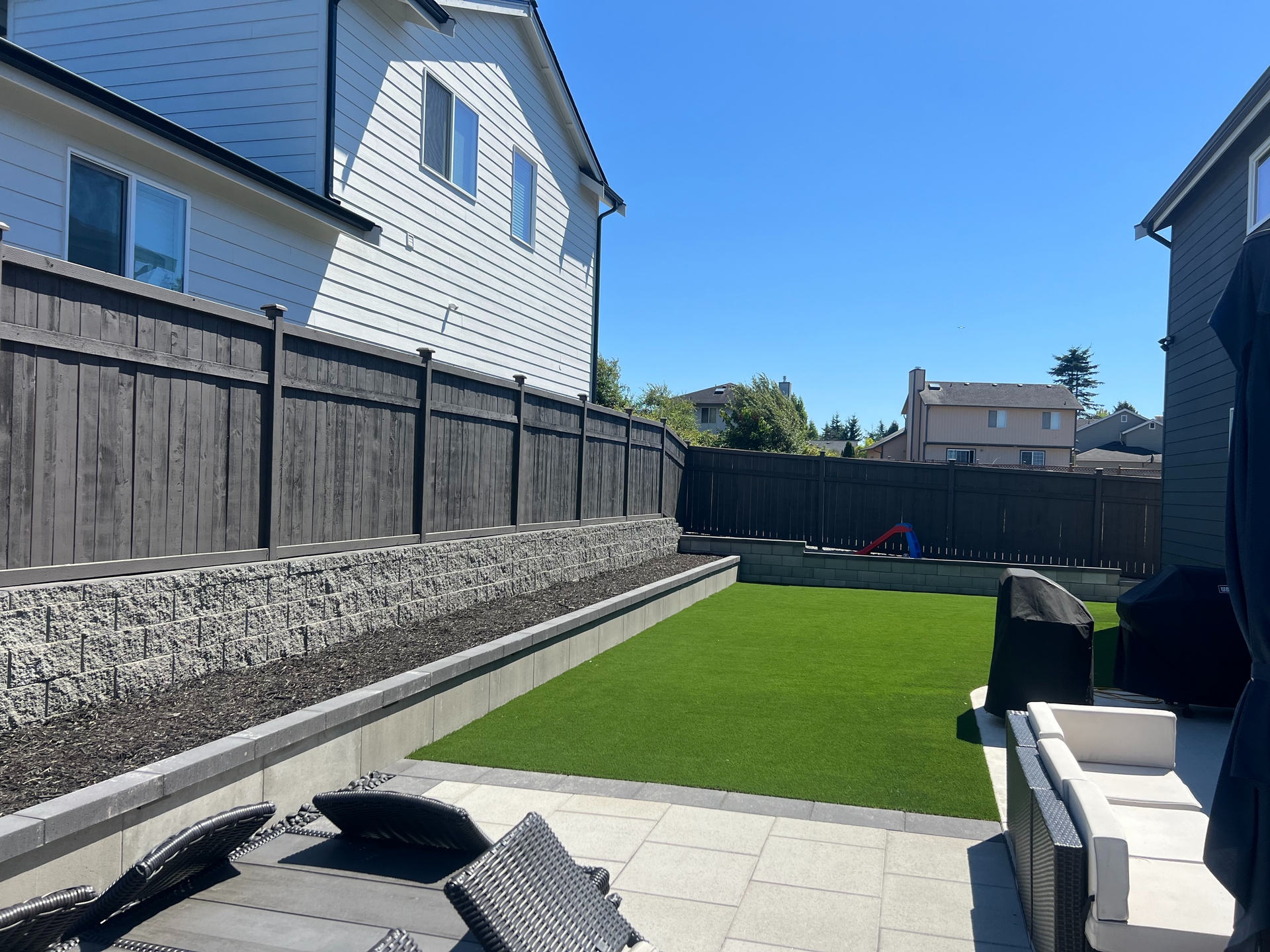 Backyard with a gray fence, retaining wall, green lawn, and patio furniture.