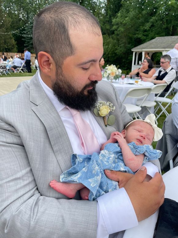 Man in a grey suit holding a sleeping baby in a blue dress. Background shows an outdoor wedding.