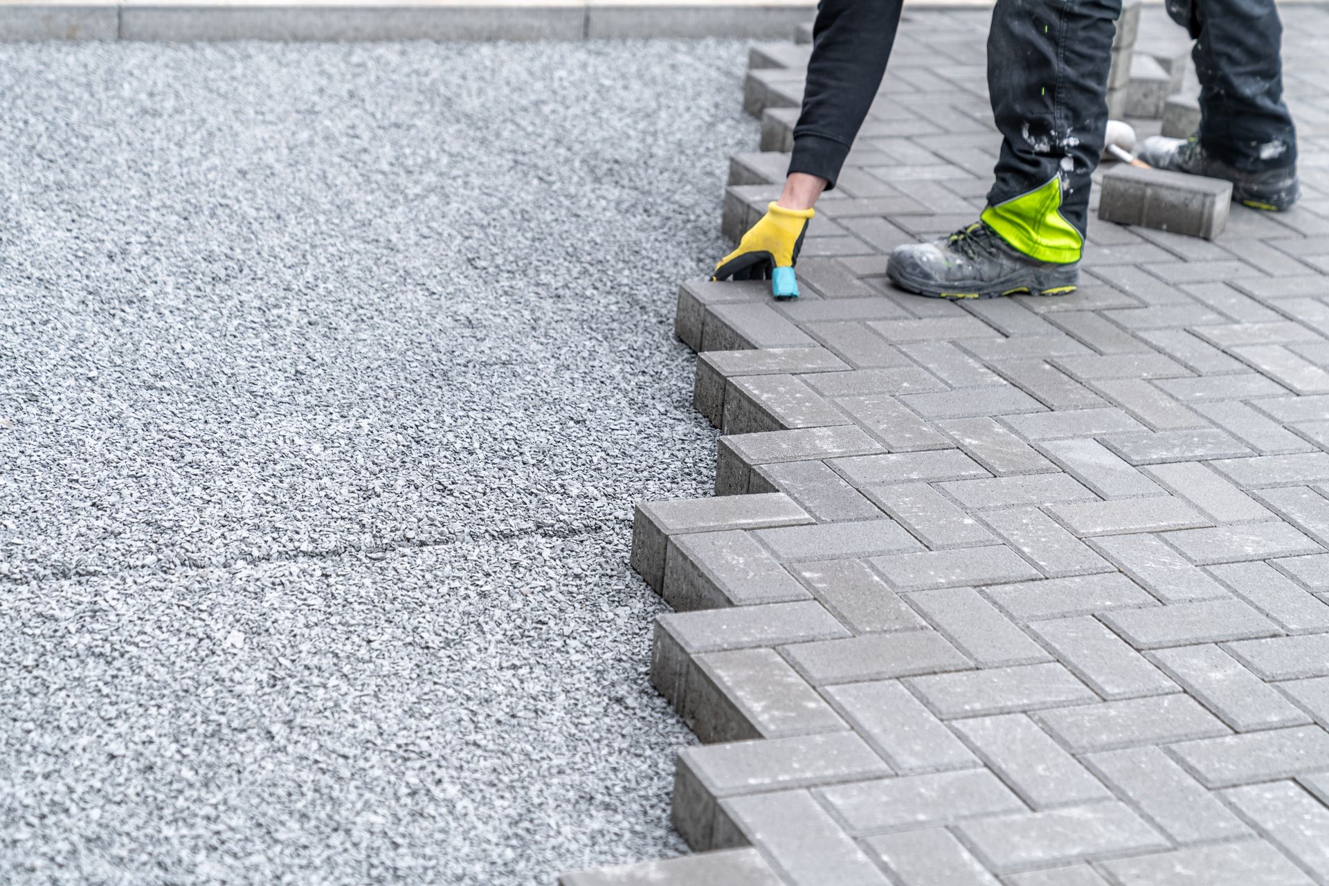 Person laying gray brick pavers on a gravel base, outdoors.