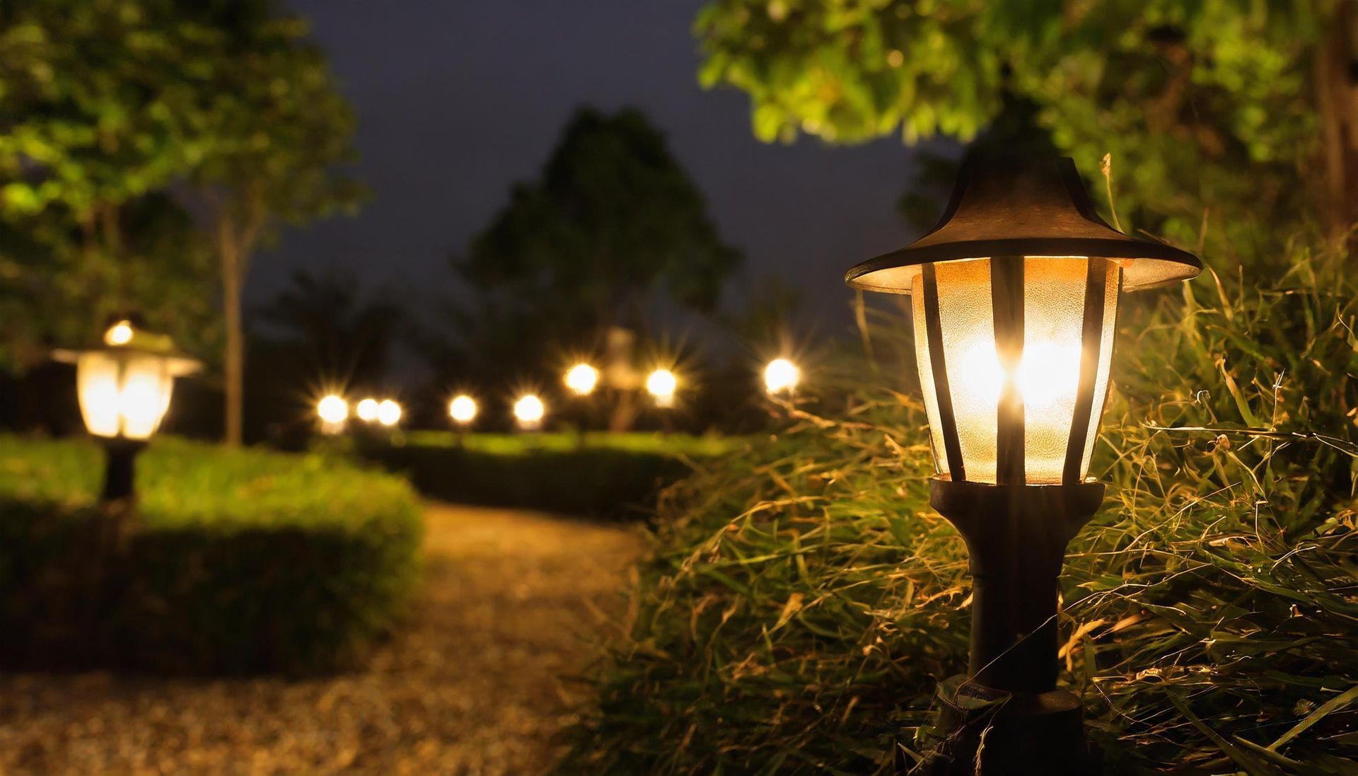 Pathway illuminated by glowing lamplights at dusk, framed by green hedges and foliage.