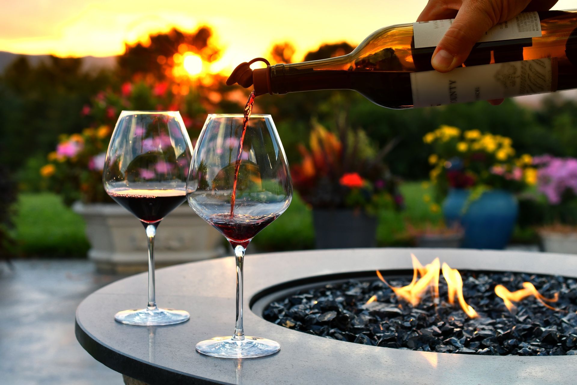 Wine being poured into glasses near a fire pit, sunset in background.