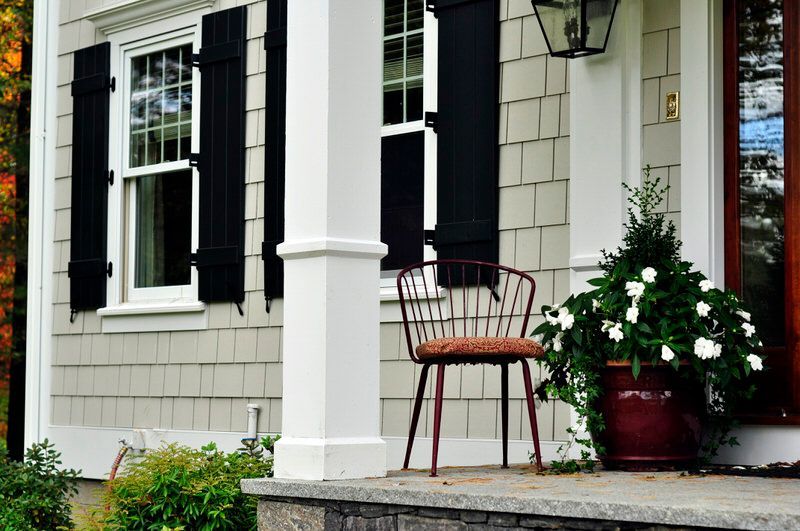 There is a chair on the porch of a house with black shutters.