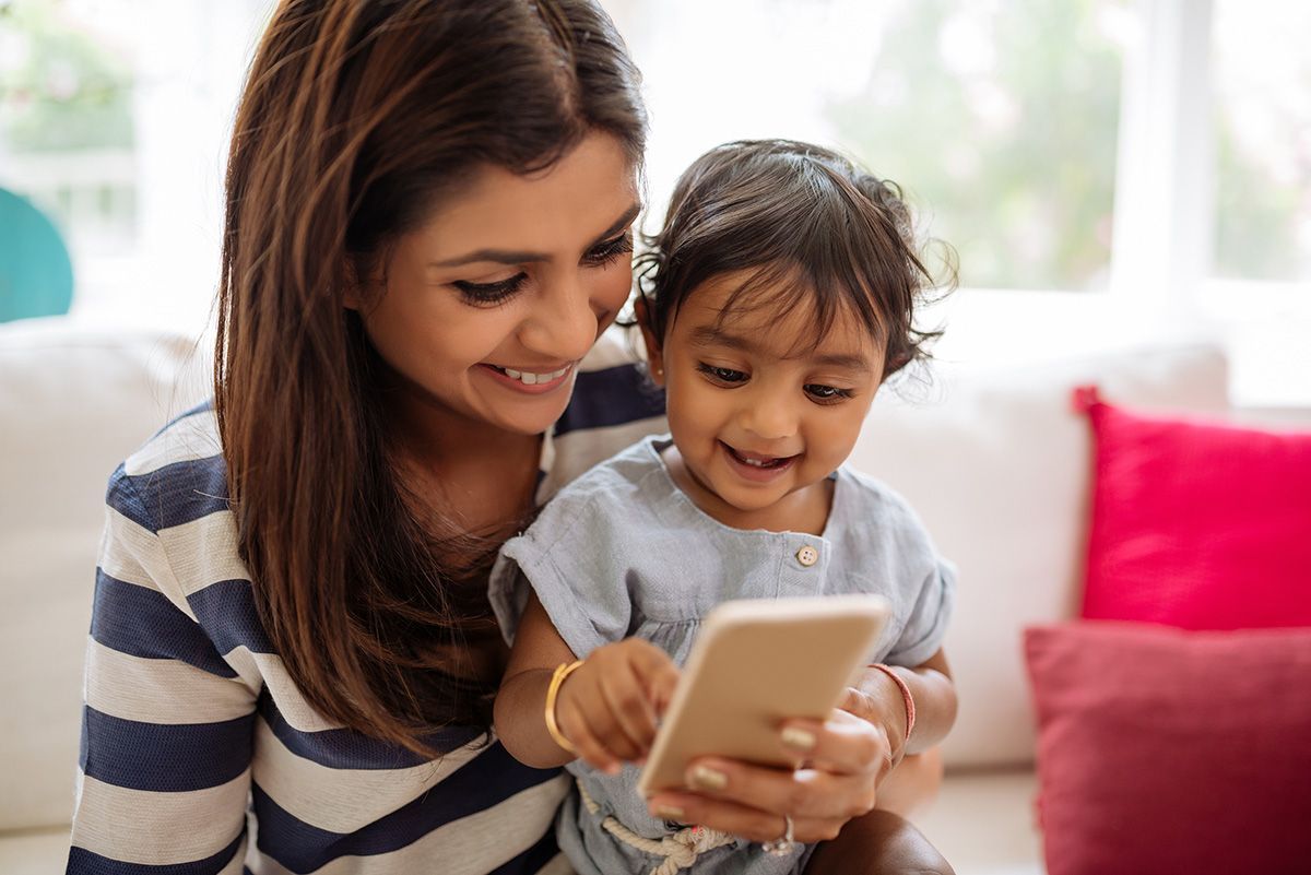 A mother is holding her child on a sofa and they are looking at a phone, smiling.