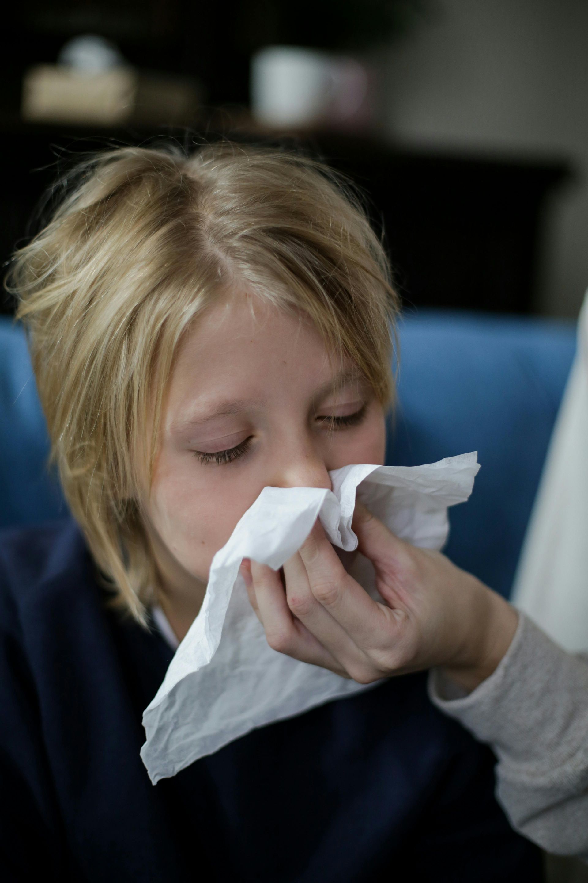 A young boy is sitting on a couch and is blowing his nose into a tissue.