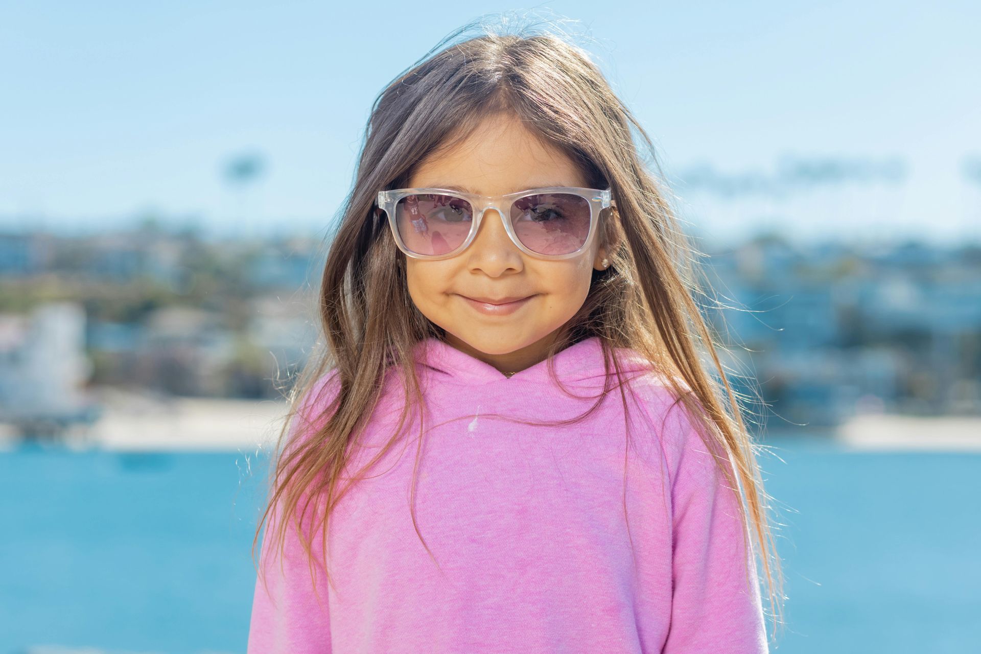 Girl wearing sunglasses and a pink hoodie smiles in front of a blue sea.