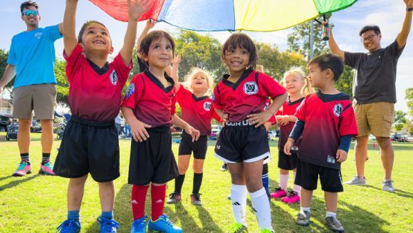 A smiling child in a blue and white sports jersey runs forward on a field, followed by another child in a red vest.