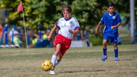A youth soccer player in a white jersey and blue shorts kicks a ball on a sunny grass field with teammates in the distance.