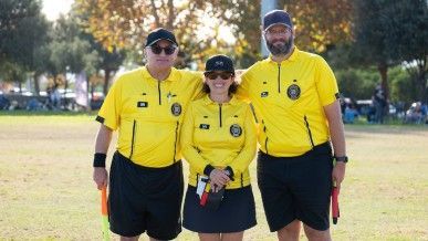 Three soccer referees in yellow shirts and black shorts stand on a field, laughing while one holds a soccer ball.
