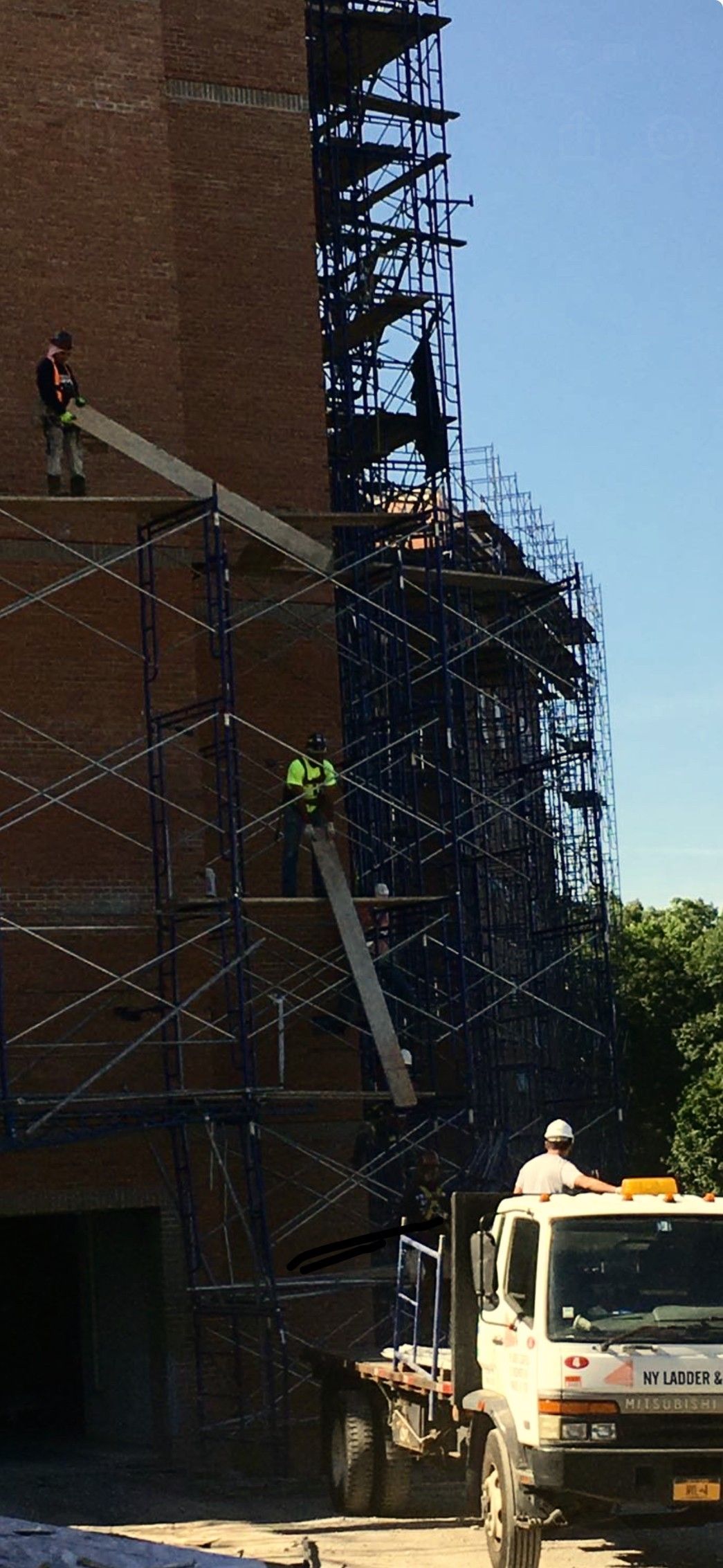 Two construction workers wearing safety harnesses and hard hats on a scaffolding, one looking at the other working.