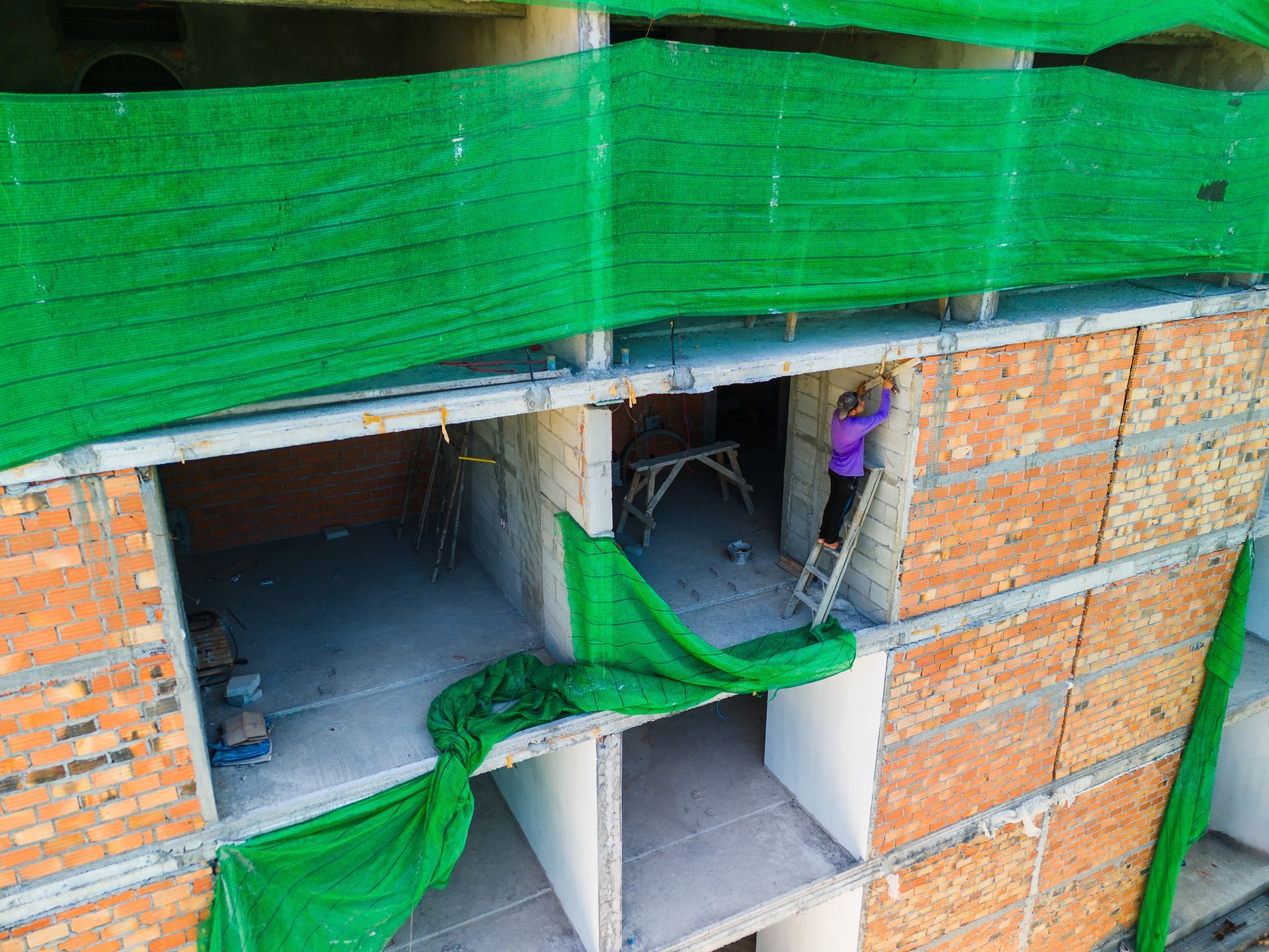 Construction worker on a ladder working on the brick facade of a building under construction, with green safety netting.