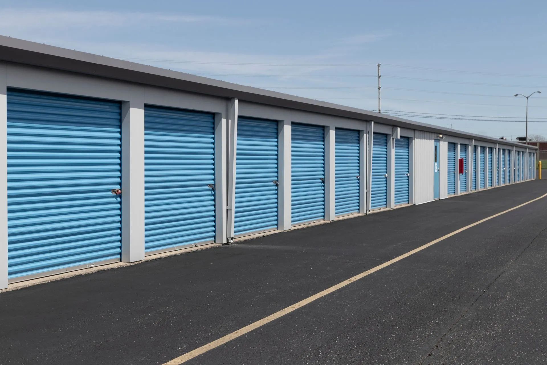 Row of storage units with blue doors and gray roof, along a paved road under a blue sky.