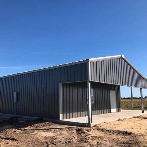 A modern, charcoal-gray metal barn with a front porch sits on a dirt lot under a clear blue sky.