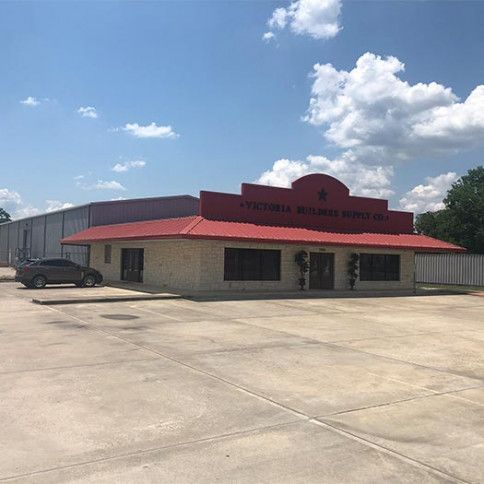 A tan, single-story building with a red roof and a large, arched facade under a bright blue sky with scattered clouds.