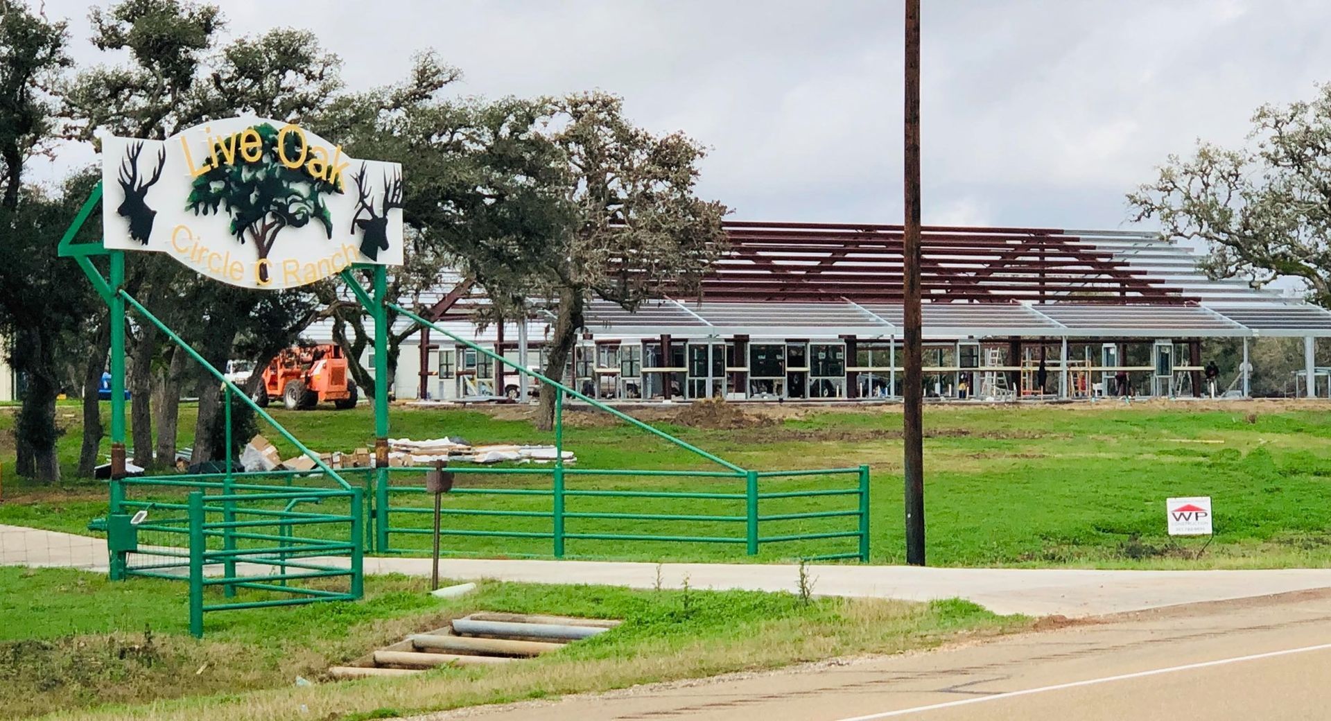A Live Oak-branded sign stands before a green field and a large metal-framed building under construction.