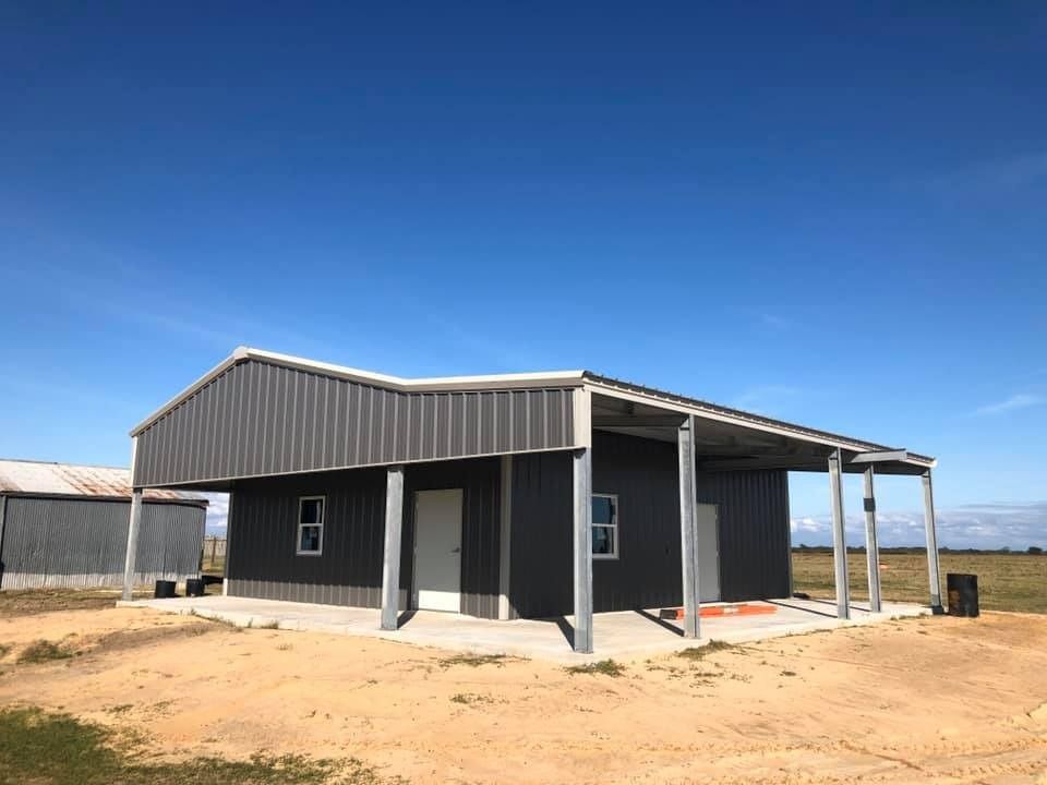 A modern dark grey metal-sided building with a covered porch and overhanging roof sits on a patch of dirt against a blue sky.