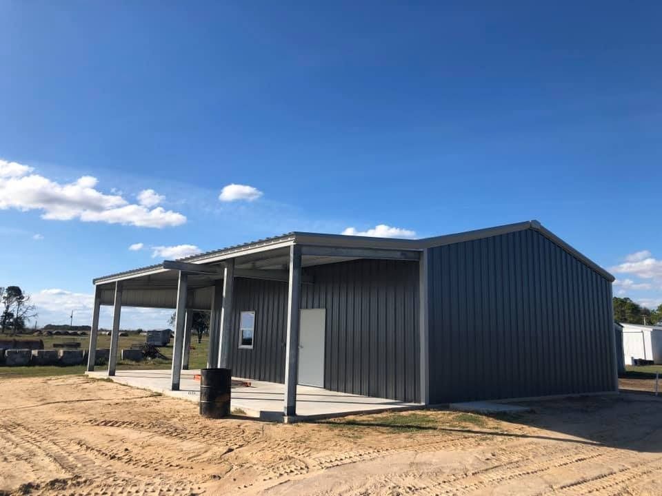 A dark gray metal building with a side covered porch, situated on a dirt lot under a bright blue sky.