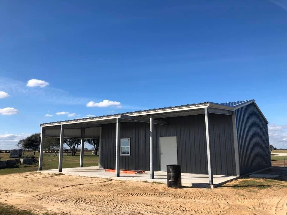 A modern, dark grey metal building with a covered porch and a white door, set in an open, grassy landscape under a blue sky.