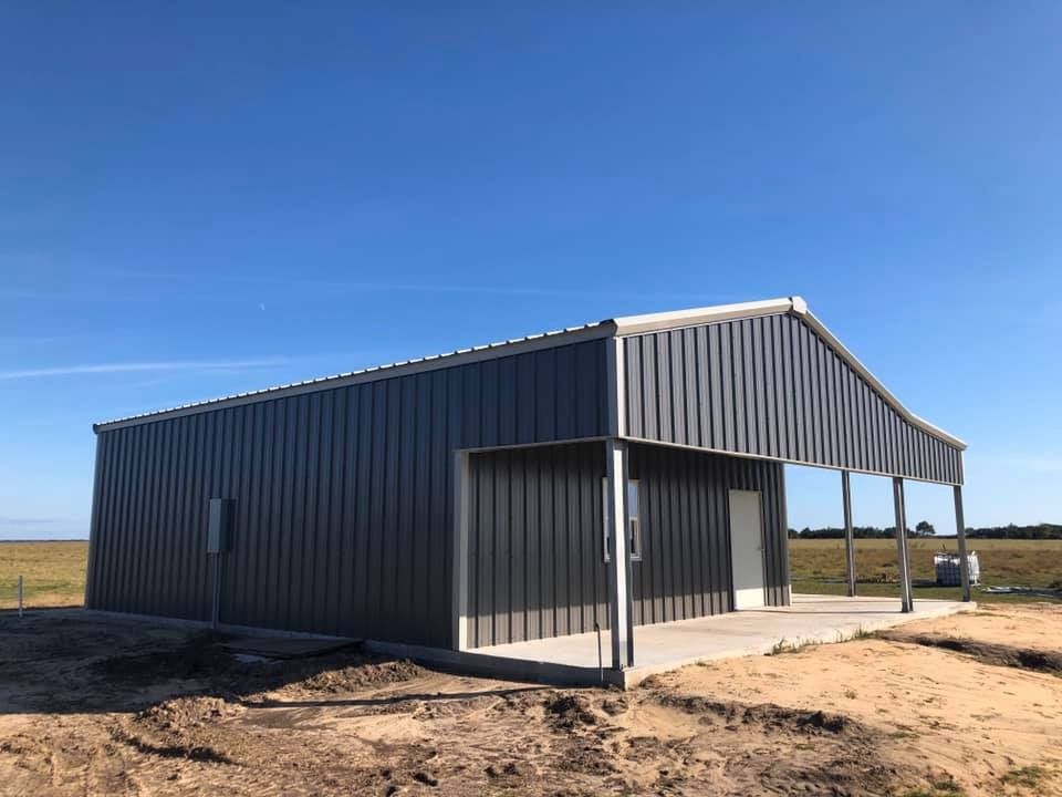 A dark grey, rectangular metal building with a side porch on a flat, grassy landscape under a clear blue sky.