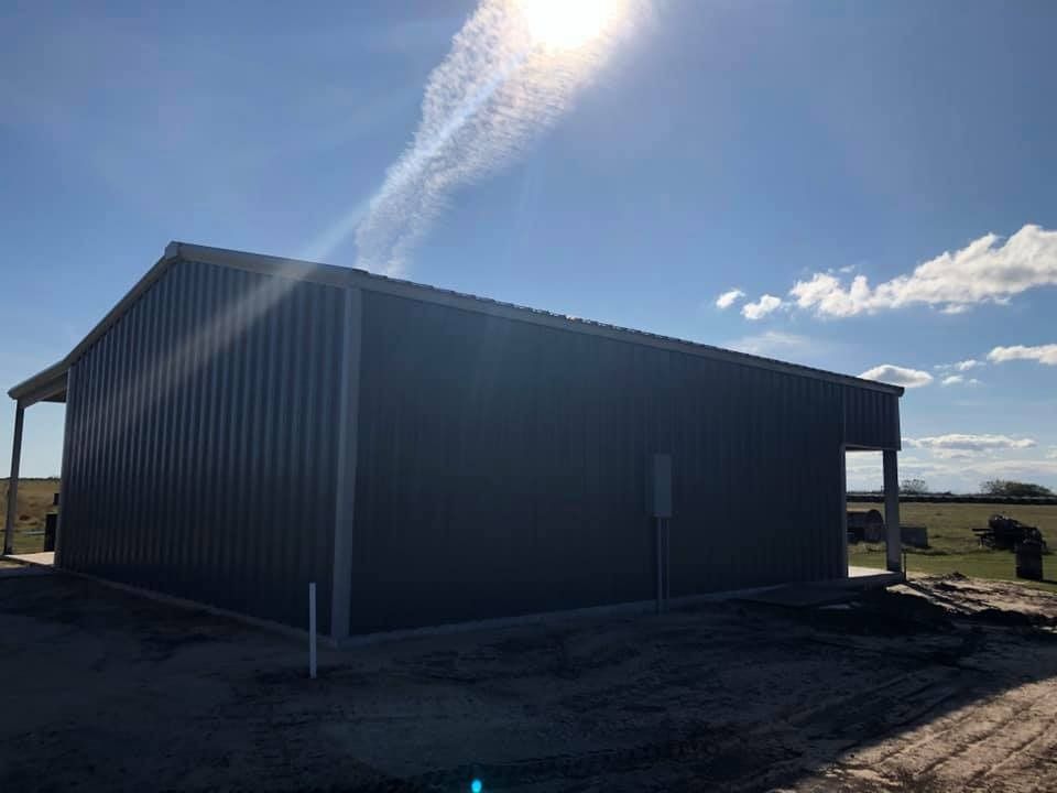 A dark gray metal building stands under a bright, sunny sky with a lens flare visible.