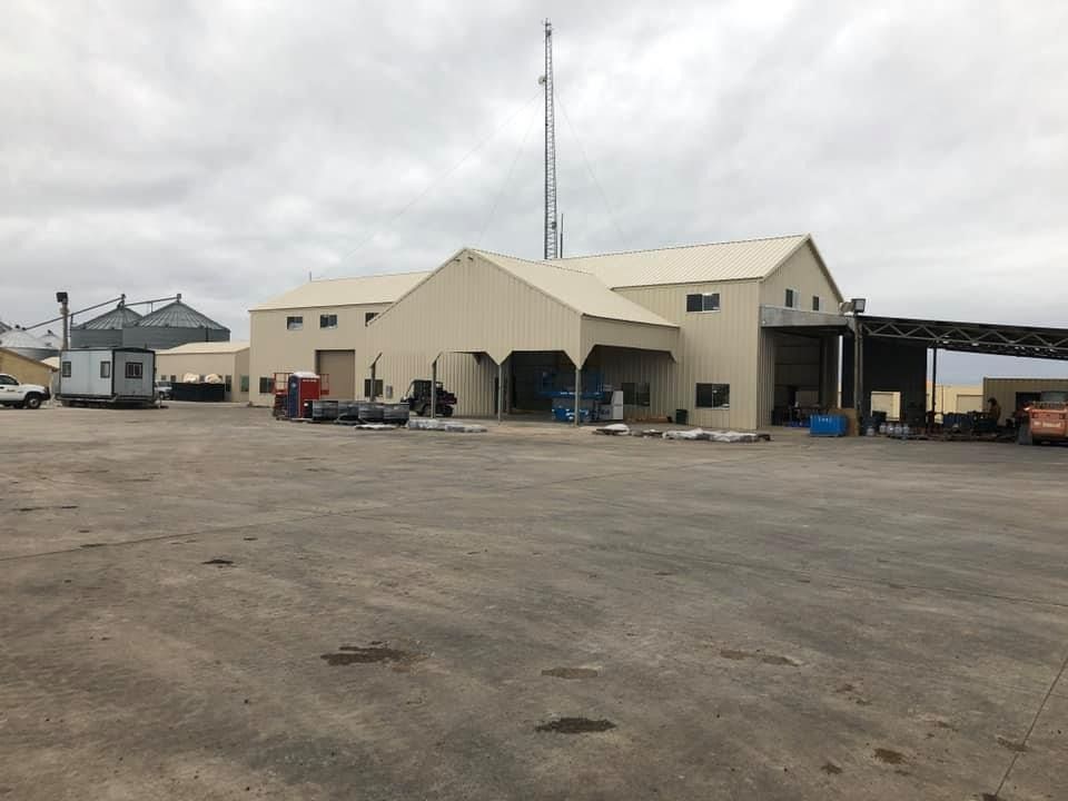 A beige industrial building with a peaked roof and a tall antenna sits on a large, flat, gravel-covered lot.