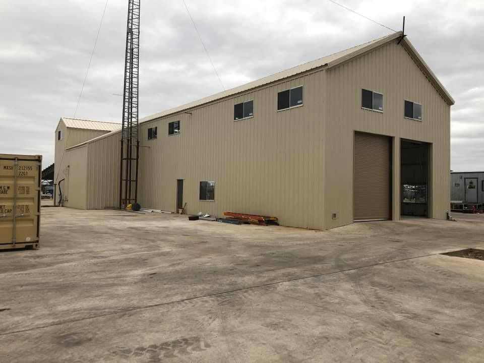 A tan metal warehouse building with two garage doors, a tall communication tower, and a gravel lot.